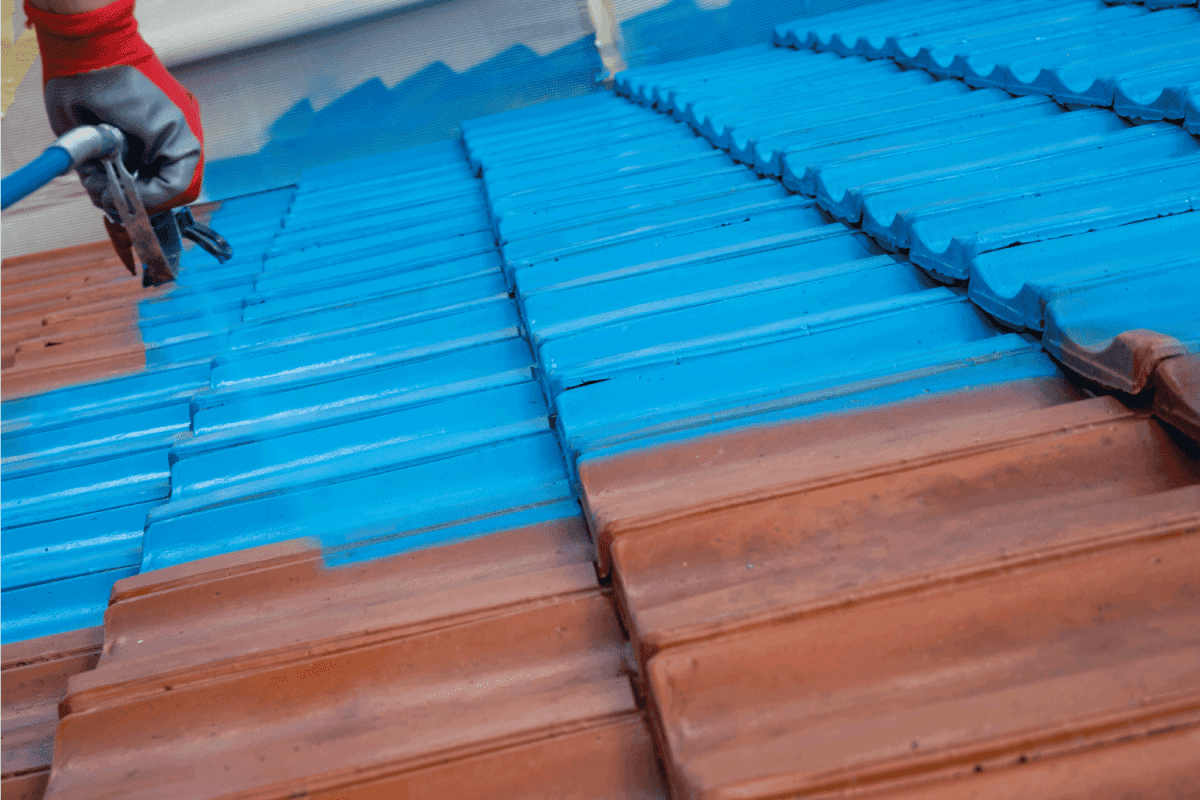 Half roof painted in blue, half of tiles are still red. worker spraying paint on terracotta roof tiles
