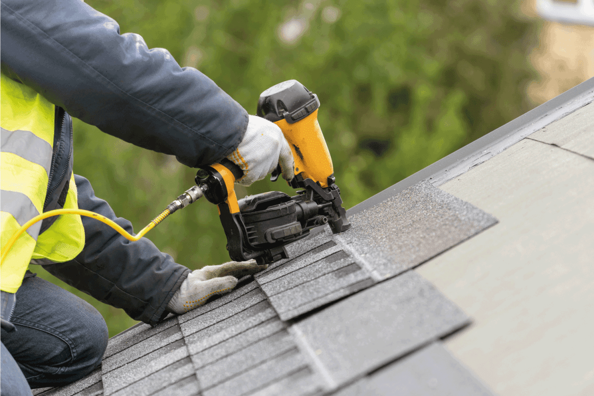 roofer worker in uniform work wear using air or pneumatic nail gun and installing asphalt or bitumen tile on top of the roof