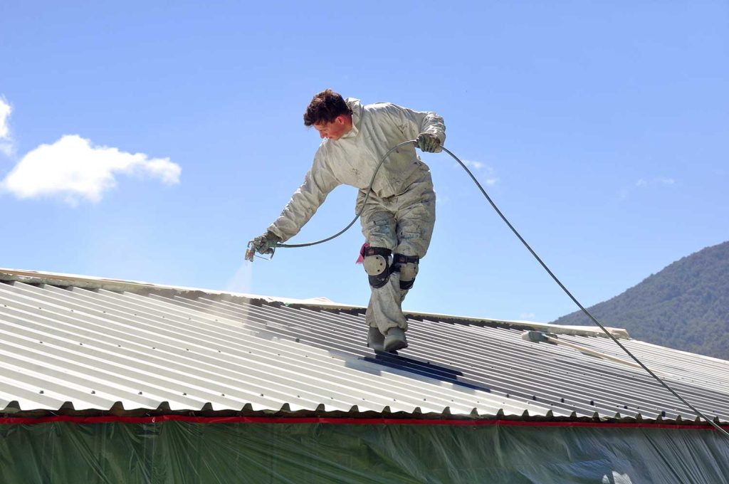 A worker uses an airless spray to paint the roof of a building