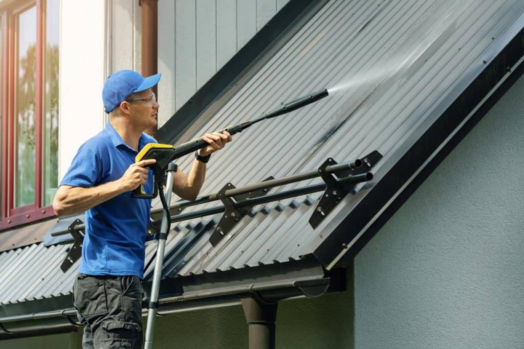 Man standing on ladder and cleaning house metal roof with high pressure washer