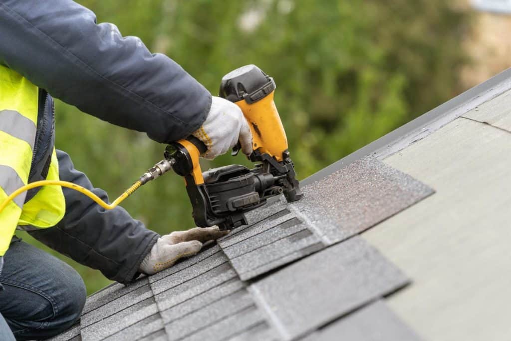 Unrecognizable roofer worker in uniform work wear using air or pneumatic nail gun and installing asphalt or bitumen tile
