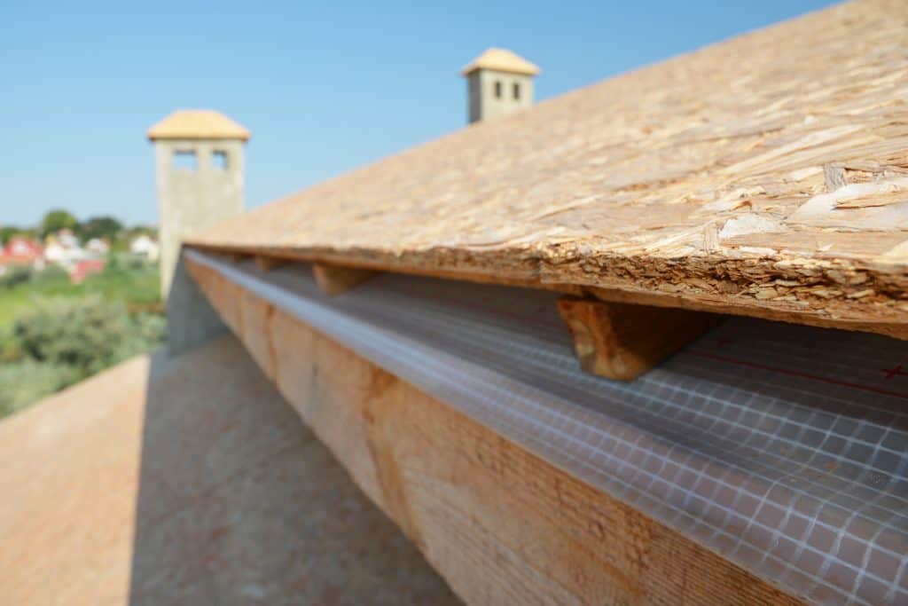 A close-up on plywood board, OSB used for roof sheathing installed on roof beams with blurred roofing construction in the background.
