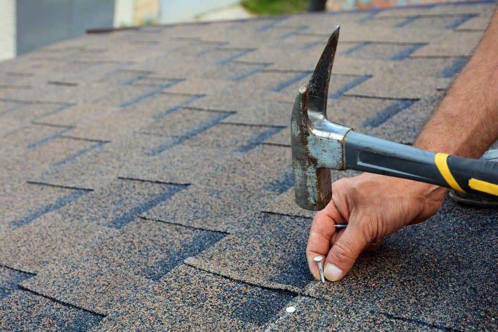 Roof cricket. Roof saddle. Worker hands installing bitumen roof shingles with hammer and nails.
